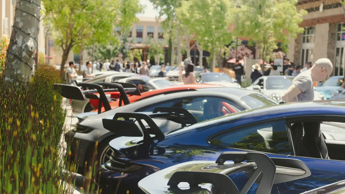 A row of Porsches with rear wings parked along a tree-lined plaza at a cars and coffee meet, with enthusiasts gathered in the background.